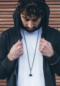 Black zip-up hoodie over a white t-shirt, with hands gripping the hoodie. A long black necklace features a small pendant. Smooth skin texture.