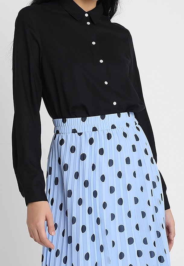 Blouse noire à manches longues avec col et boutons blancs, associée à une jupe plissée bleu clair ornée de gros pois noirs.
