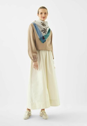 Vrouw glimlachend, draagt een beige blouse, crèmekleurige broek met wijde pijpen, een blauw-groen patroon sjaal en witte sneakers, staand tegen een witte achtergrond.