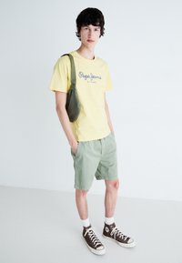 Geltonas medvilninis marškinėliai su "Pepe Jeans" logotipu, žali šortai, rudi aukšti sportbačiai ir žalia peties rankinė. Paprasta fonas.