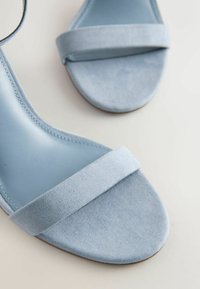Paire de sandales en daim bleu clair à bout ouvert avec une bride unique à l'avant, vues de dessus sur un fond clair.