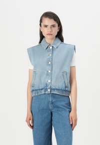 Colete de ganga sem mangas com fecho de botão, colarinho e bainha elástica. Lavagem azul clara com bolsos frontais e detalhes de costura visíveis.