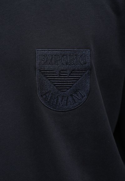 Mørkeblått stoff med en brodert merkepute med "EMPORIO ARMANI" og en stilisert ørnlogo, tydelig vist på brystet.