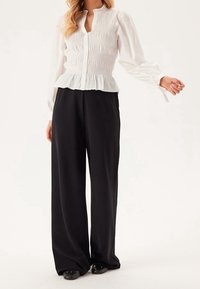 Weißes, gerafftes Oberteil mit langen Ärmeln und Rüschensaum, kombiniert mit hochgeschnittenen, schwarzen Schlaghosen. Schwarze flache Schuhe runden das Outfit ab.