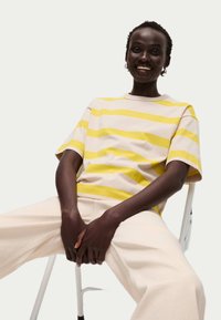 Pruhovaný tričko v světlé béžové barvě s výraznými žlutými pruhy, kulatý výstřih, volný střih. Model usazený v krémových kalhotách s širokými nohavicemi na neutrálním pozadí.