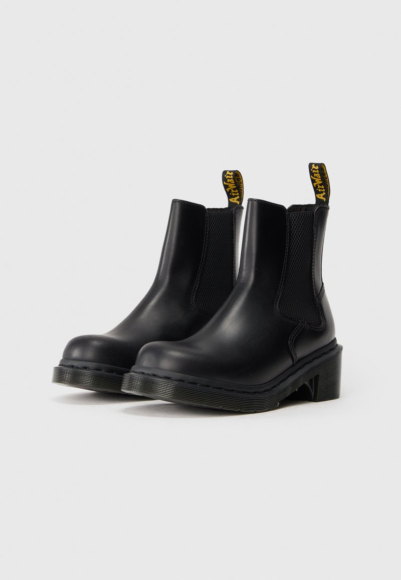 Par de botines de cuero negro con paneles laterales elásticos y bucles de talón amarillos con marca, sobre fondo blanco.