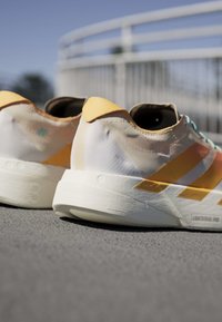 Close-up van witte en gele lichte hardloopschoenen op asfalt met een wazige metalen reling op de achtergrond onder een heldere hemel.
