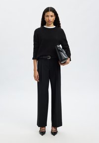 Svart stickad tröja, svarta byxor med vida ben och svarta spetsiga klackar. Håller i en strukturerad svart handväska med ett vikbart övre fack.