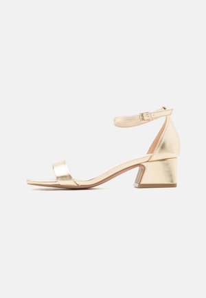 Gold ankle-strap sandal with a low block heel and open toe on a white background.