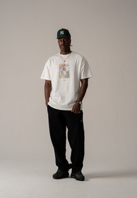 Hvit bomull T-skjorte med grafisk trykk, løse sorte bukser, grønn baseballcap, lagdelte kjeder og mørke sneakers.