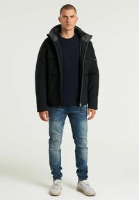 Chaqueta negra con capucha, cierre frontal, múltiples bolsillos y forro gris. Llevada sobre una camiseta azul marino, combinada con vaqueros azules desgastados y zapatillas.