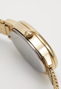 Montre dorée avec un boîtier rond, une couronne texturée et un bracelet en métal avec un design à maillons. Le dos a une surface lisse.