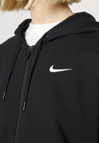 Svart zip-up hoodie i mjukt tyg, med en vit Nike-logotyp på vänster bröst och justerbara dragkedjor på huvan.