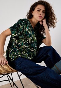 Kurzärmlige Bluse in grün, schwarz und weißem abstraktem Muster; lockere Passform mit V-Ausschnitt, kombiniert mit dunkelblauen weiten Jeans.