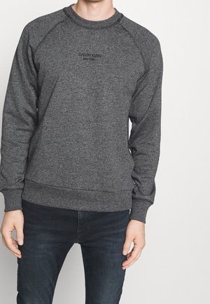 Mand iført mørkegrå Calvin Klein sweatshirt med sort tekst og mørke jeans, stående mod en ensartet lys baggrund.