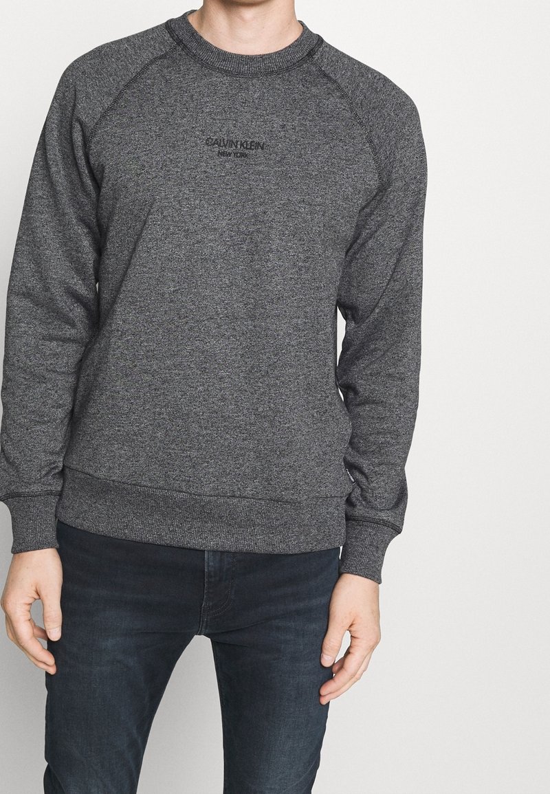 Homme portant un sweat-shirt Calvin Klein gris foncé avec un texte noir et un jean foncé, debout devant un fond clair uni.