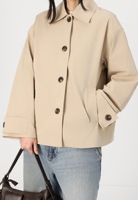 Beige, oversized jakke lavet af bomuldsblanding, med spidst krave, store brune knapper og to sidelommer. Bæres med denimjeans.