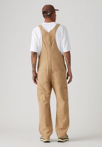 Salopette en coton beige avec une coupe ample, dotée de poches avant et latérales, de bretelles réglables et d'un empiècement au dos. Portée par-dessus une chemise blanche.