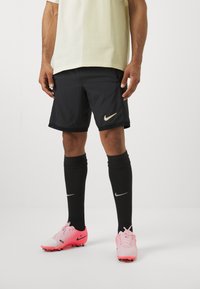Shorts de sport noirs avec un logo Nike doré, associés à des chaussettes noires montantes et des crampons roses avec des accents blancs et noirs.