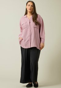 Camisa de color rosa claro, con cuello, dos bolsillos en el pecho, botones al frente y mangas largas. Combinada con pantalones anchos negros y mocasines negros.