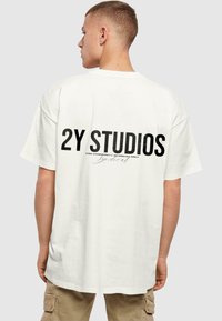 Valge puuvillane T-särk üle mõõtu sobivuses, mille seljal on julge must tekst "2Y STUDIOS" ja väiksem tekst all. Lühikesed varrukad, ümmargune kaelus.
