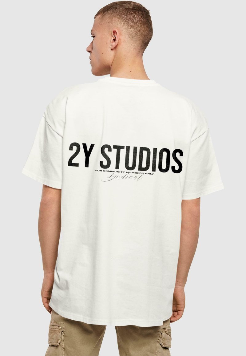Valge puuvillane T-särk üle mõõtu sobivuses, mille seljal on julge must tekst "2Y STUDIOS" ja väiksem tekst all. Lühikesed varrukad, ümmargune kaelus.