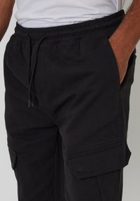 Schwarze Cargo-Hosen mit elastischem Bund, Seitentaschen und einer glatten, matten Textur. Verfügt über doppelt genähte Nähte für Langlebigkeit.