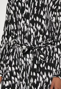 Schwarzes Kleid mit einem weißen abstrakten Pinselstrichmuster, das eine Taillenbindung und lange Ärmel aufweist. Der Stoff wirkt glatt und hat eine weiche Textur.