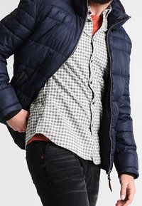 Chaqueta acolchada azul marino con un acabado texturizado, llevada sobre una camisa gris a cuadros. Presenta un cuello alzado y cierre con cremallera. Jeans negros completan el conjunto.