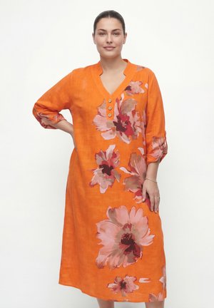 Woman wearing a bright orange knee-length dress with large pink and red floral patterns, three buttons at the neckline, and rolled-up sleeves.
