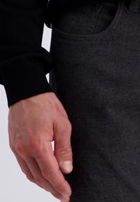 Zwarte longsleeve shirt met aansluitende mouwen en een gestructureerde donkergrijze broek. Hand rustend aan de zijkant, die de natuurlijke huidtint en -vorm toont.