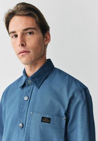 Blå skjorte med knap foran og et kordfløjlskrave, der har en brystlomme med et logo-tag og strukturerede stofdetaljer.