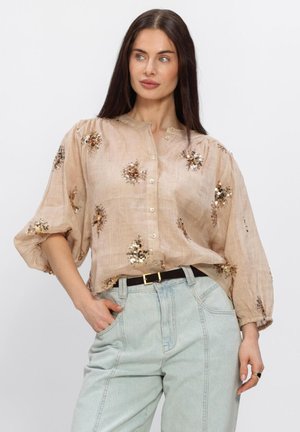Vrouw die een beige blouse met goudkleurige, met pailletten versierde bloemenpatronen draagt en lichtblauwe, hooggesneden jeans, poserend met één hand in de zak.