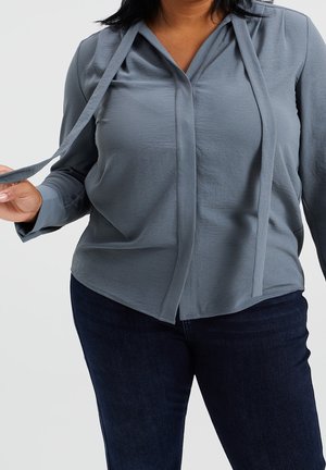 Frau trägt eine graue Langarmbluse mit herabhängenden Bändern, kombiniert mit dunkelblauen Jeans, hält ein Band in der Hand vor einem einfarbigen Hintergrund.