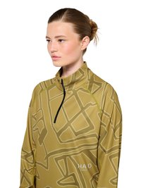 Ung kvinde med håret sat op i en knold bærer en gul trøje med høj krave med et geometrisk mønster og en sort lynlås på hvid baggrund.