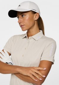 Beżowa koszulka polo z krótkimi rękawami i zapięciem na guziki, z haftowanym białym logo na kołnierzyku i rękawie. Obok biała rękawica golfowa.