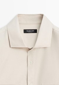 Chemise boutonnée couleur crème avec un col classique, affichant une étiquette Massimo Dutti cousue à l'intérieur de l'encolure.