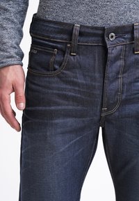 Mörkblå denimjeans med en figurnära design, guldsömmar, knappstängning och flera fickor. Tyget har en slät yta.