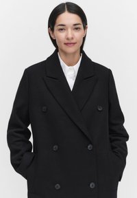 Svart dubbelknäppt kappa i tjockt tyg, med stora knappar, en spetsig krage och framfickor, bärs över en vit skjorta.