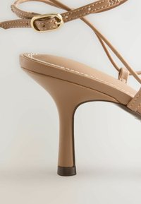 Sandale à talon haut beige avec bride à la cheville, boucle dorée, et détail de couture blanche sur la semelle rembourrée.
