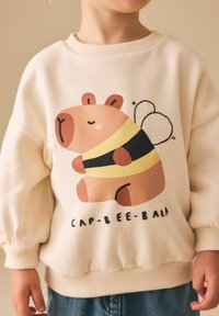 Sweat-shirt beige clair avec un dessin d'un ours de dessin animé avec des ailes d'abeille, un t-shirt rayé jaune et noir, et le texte "CAP-BEE-BARA" en dessous.
