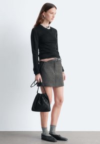 Schwarzes langärmliges Oberteil, grauer Rock mit Gürtel, schwarzer Beuteltasche mit Kordelzug, graue Socken, schwarze Slipper. Einfaches, figurbetontes, strukturiertes Outfit.