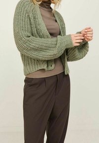 Groene gebreide cardigan met een textuurpatroon, open voorkant en lange mouwen, gecombineerd met een bruine top en donkere broeken. Soepele stof en ontspannen pasvorm.