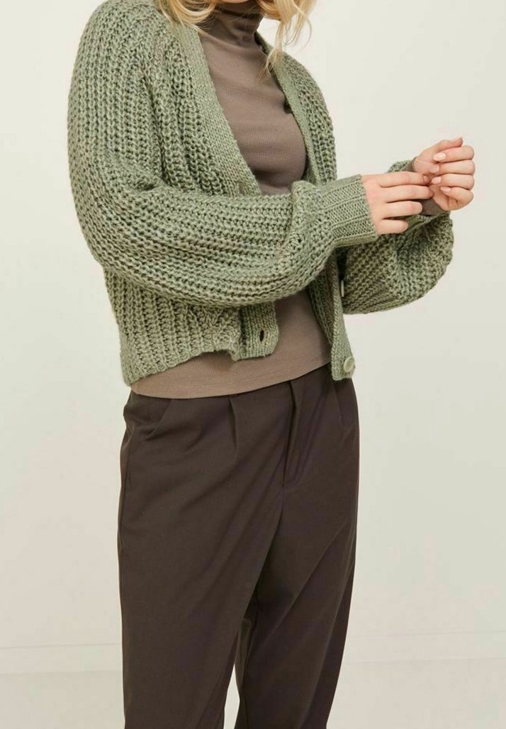 Groene gebreide cardigan met een textuurpatroon, open voorkant en lange mouwen, gecombineerd met een bruine top en donkere broeken. Soepele stof en ontspannen pasvorm.