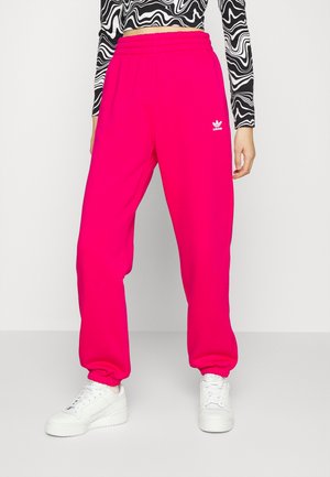 Osoba nosząca jasnoróżowe spodnie dresowe Adidas, czarno-białą, wzorzystą długą bluzkę typu crop top oraz białe tenisówki, stojąca na jednolitym tle.