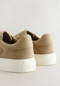 Paire de baskets basses en daim beige avec semelles épaisses en caoutchouc blanc, vues de dos sur un fond blanc uni.