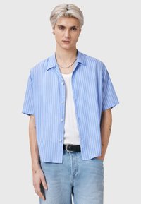 Camicia azzurra chiara a maniche corte con righe bianche verticali, chiusura con bottoni sul davanti e una texture liscia. Indossata sopra un top bianco senza maniche.