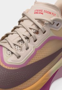 Κοντινή λήψη από τρέξιμο παπούτσι Nike Zoom Fly σε μπεζ και μοβ χρώμα, με λεπτομερείς κορδόνια και επάνω μέρος από δίχτυ, δείχνοντας το λογότυπο στη γλώσσα.