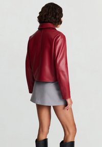 Veste courte en cuir rouge avec un col large et des manches longues, présentant une texture lisse. Associée à une mini-jupe grise et des bottes noires montantes.