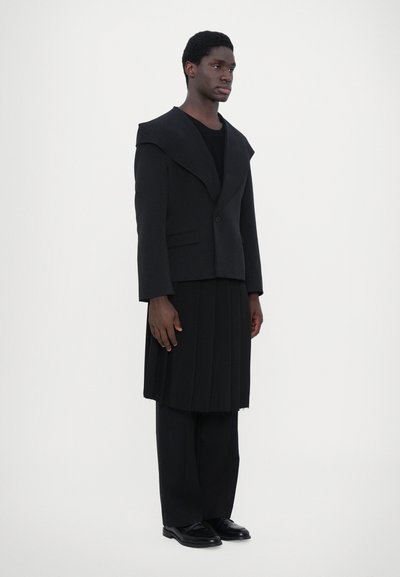 Schwarze taillierte Jacke mit breiten Revers, kombiniert mit einem plissierten schwarzen Rock und gestreiften Hosen. Das Modell steht vor einem schlichten weißen Hintergrund.